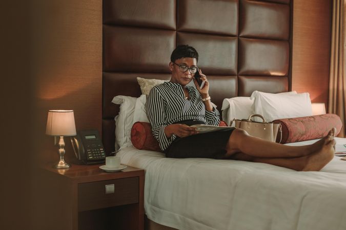 Woman in formal clothes sitting on bed talking on mobile phone with newspaper in hand