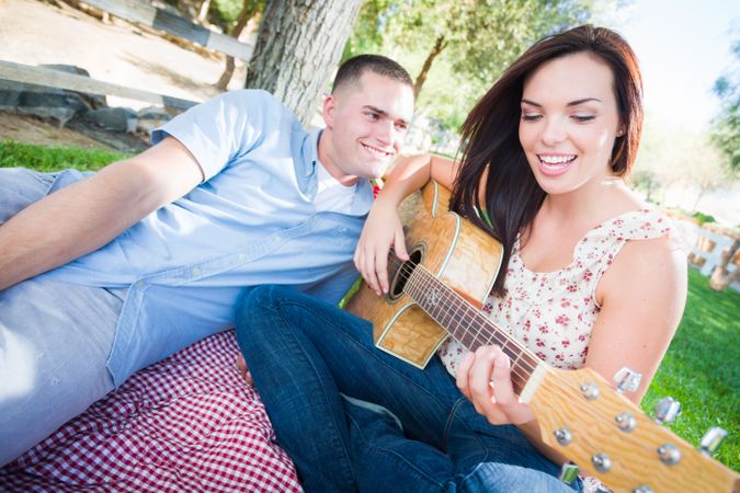 Young Adult Playing Guitar with Boyfriend In The Park.