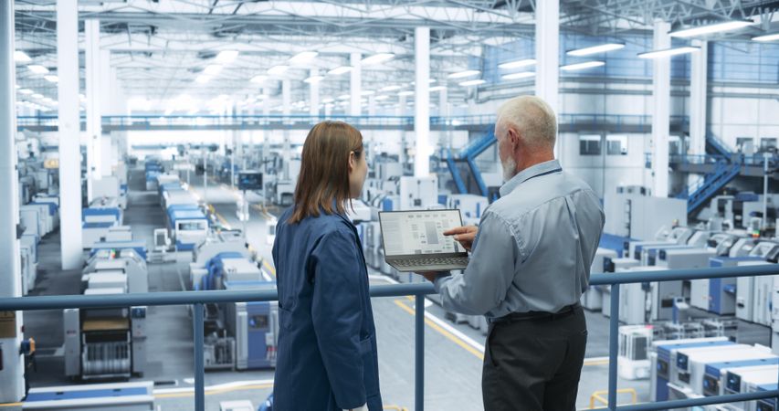 Two Multiethnic Engineers Standing on a Platform, Using Laptop Computer at an Electronics Factory. Machines are Undergoing Maintenance, Specialists Monitoring the Progress Through Online Software
