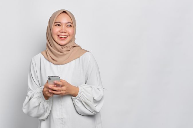 Asian female in headscarf looking happy while looking up from cell phone