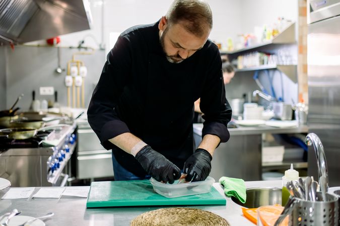 Bearded male professional chef in black gloves arraigning food into transparent container while working in professional kitchen