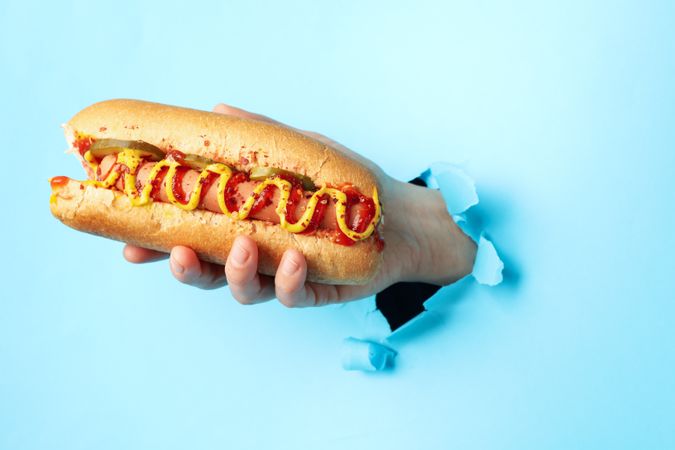 Hand holds hot dog from hole on blue background