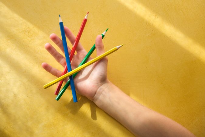 A child's hand picking up coloured pencils on a yellow backgroun