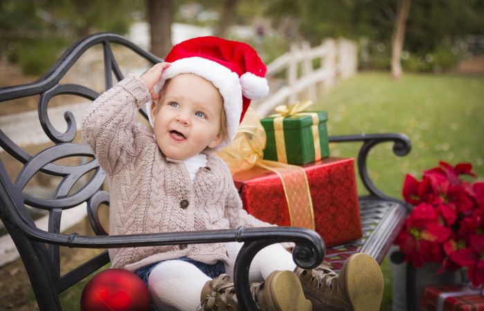 Young Child Wearing Santa Hat Sitting with Christmas Gifts Outside.