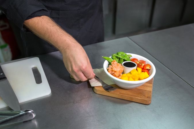 Unrecognizable chef putting fork next to fresh colorful poke bowl with salmon on restaurant kitchen