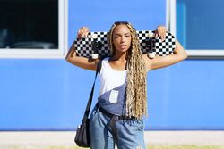 Female in denim overalls standing with skateboard behind head in front of blue building