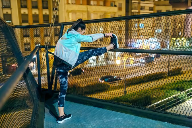 Unrecognizable brunette woman athlete stretching her legs over bridge railing before run at night