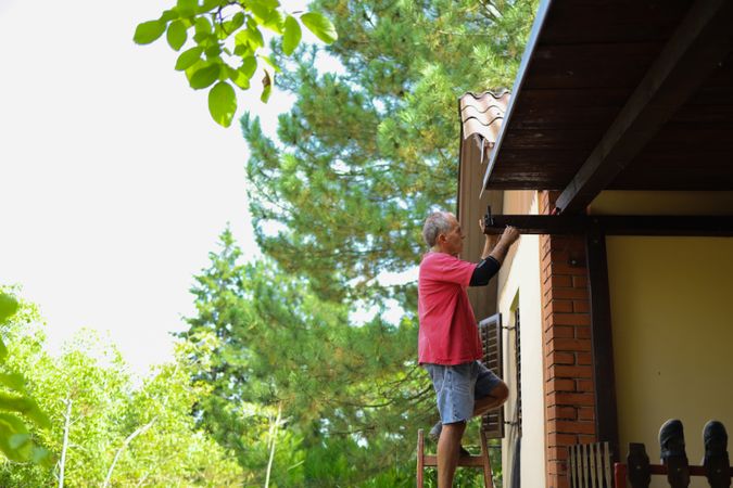 Roofline Repairs: Older Man on Ladder Fixing Wooden Beam of House Exterior.