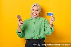 Happy Muslim woman in headscarf and green blouse holding credit card and smart phone up bGw8Yb