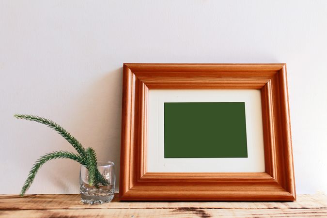 Rectangular wooden picture frame leaning against wall next to branch in glass mockup