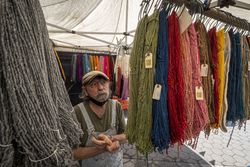 Man with overalls and cap stands near brightly colored sheep wool at a farmers market 4B6KM4