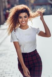 Happy Arab woman with curly hair walking down sunny street playing with hair