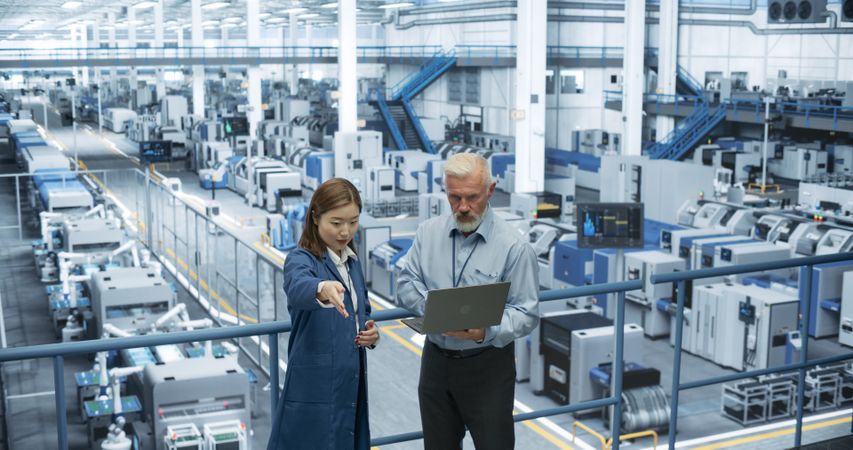 Portrait of a Mature Engineer and Happy Asian Scientist Using Laptop Computer and Talking in a Factory Facility with Equipment Producing Modern Electronic Components for Different Industries