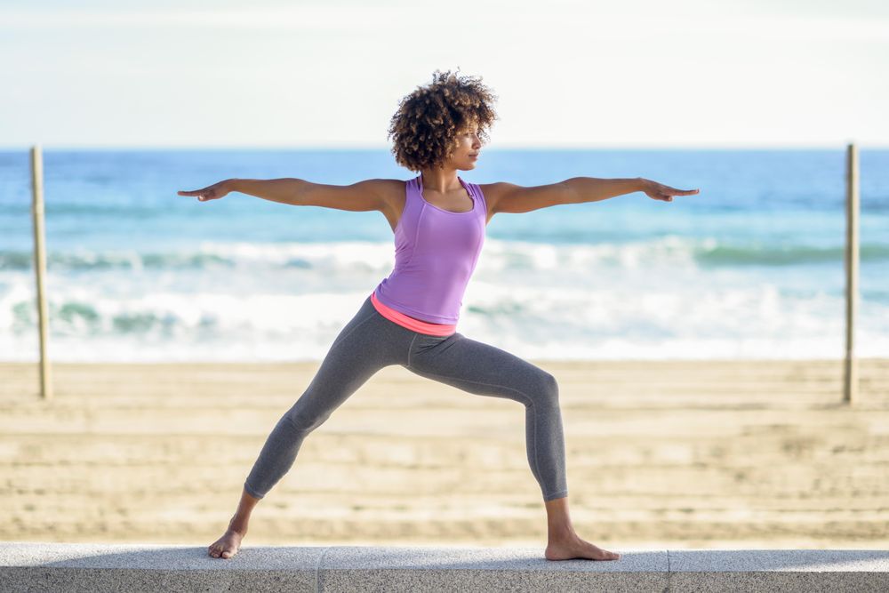 Black woman with afro hairstyle doing yoga warrior pose near beach