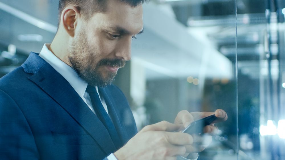 Businessman Standing in the Office Hallway Uses Smartphone, Doing Gestures.