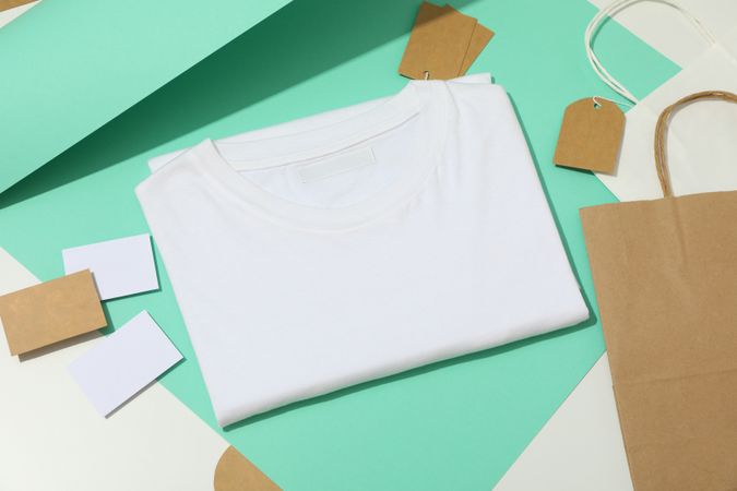 Blank t-shirt, tags, paper cup and paper bags on mint and light background, space for text