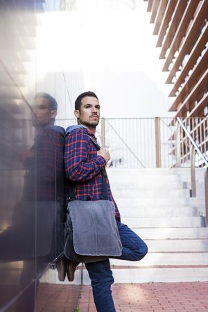 Man in plaid shirt leaning on wall outdoors in front of stairs on a sunny day, vertical