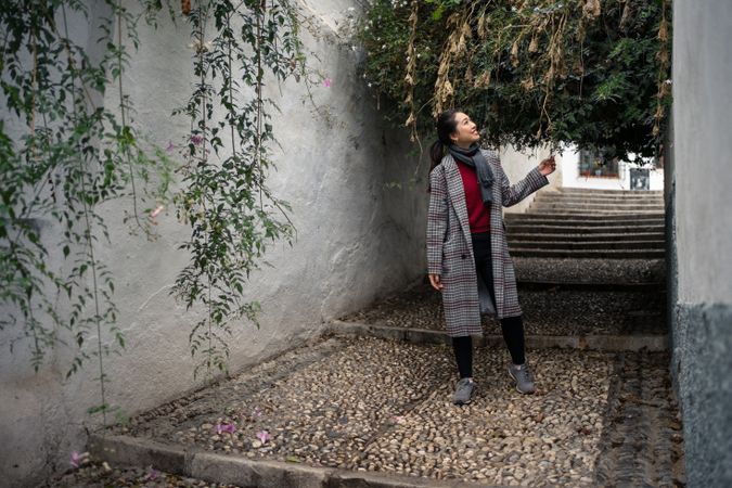 Young female in casual clothing touching green plants and smiling at rocked lane