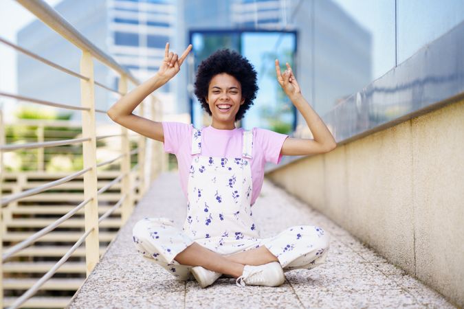 Smiling woman in floral overalls sitting cross legged with hands up