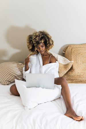 Black woman drinking coffee while sitting on bed with laptop