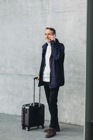 Older male tourist standing outside with suitcase on phone, vertical