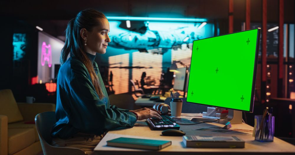 Portrait of Young Woman Sitting at Her Desk Using Desktop Computer with Mock-up Green Screen.