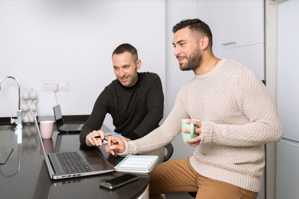 Two men sitting at kitchen counter and taking notes from laptop screen