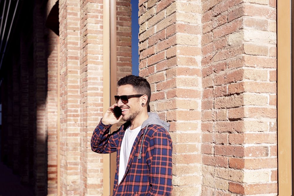 Bearded man leaning on a brick wall wearing sunglasses while using a smartphone in the sun