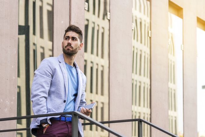 Portrait of a happy bearded young man holding a mobile phone while standing outdoors and looking away