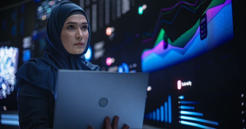 Muslim Female Data Center IT Engineer Standing in a Room with Statistics and Charts Settings on a Digital Screen.