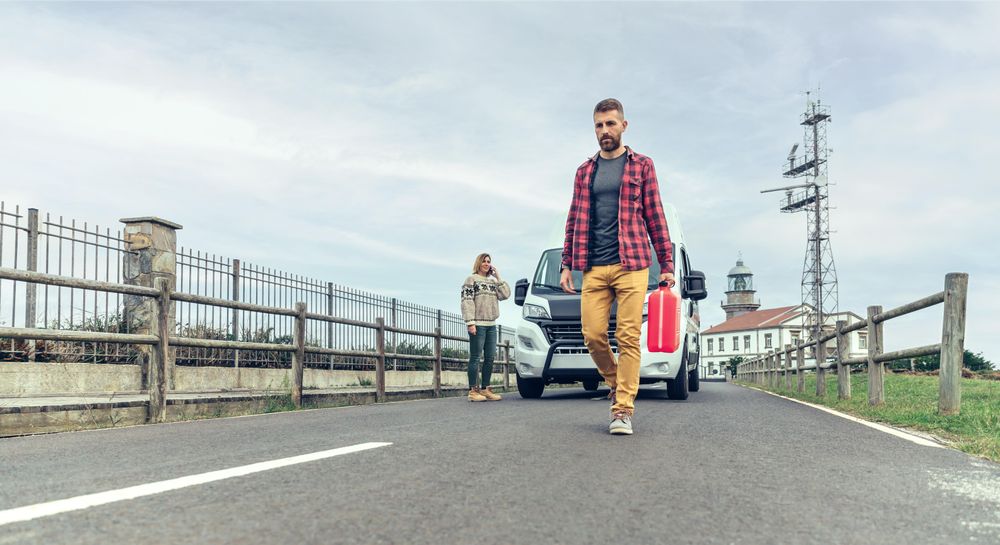 Male in red checkered shirt walking in front of broken down van with gas can