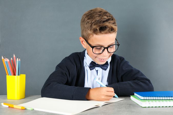 Boy in school uniform writes sitting at the table against board background