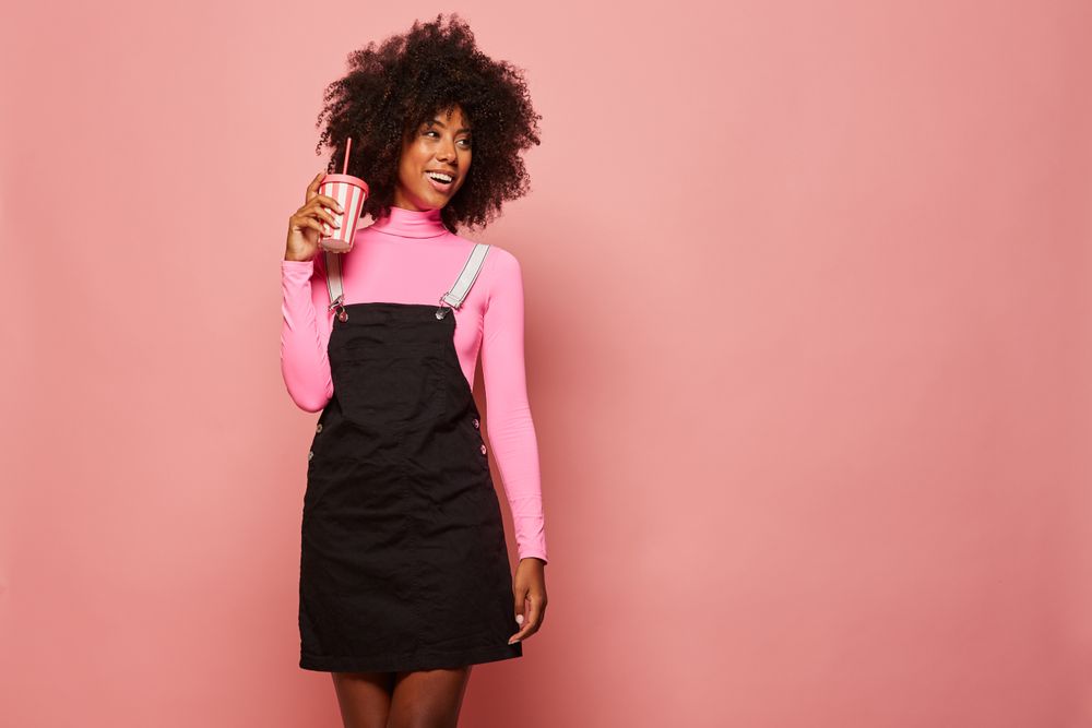 Happy Black woman with disposable cup of drink standing against pink background