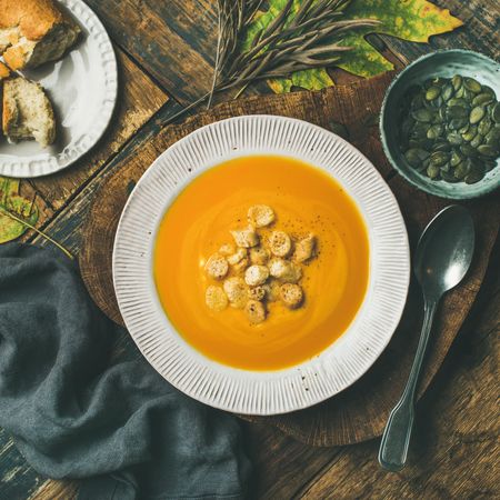 Pumpkin soup with croutons, bread, seeds, spoon on wooden table with leaves, square crop