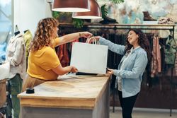 Fashion store owner handing over shopping bag to female customer at checkout counter