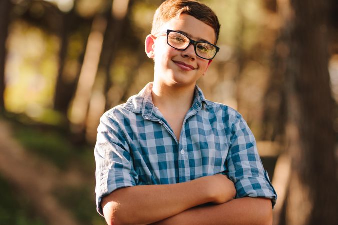 Smiling boy in eyeglasses standing in a park with arms crossed