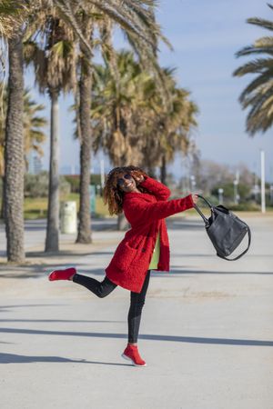 Smiling female in sunglasses with curly hair jumping on palm tree lined street