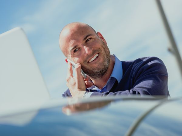 Businessman talking on the phone while using the laptop supported by a car and smiling expression
