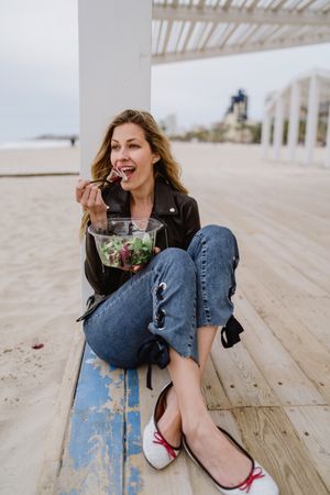 Content woman eating salad from plastic bowl sitting on wooden terrace