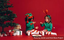 Little girls sitting beside a Christmas tree enjoying cookies and milk 47JGP5