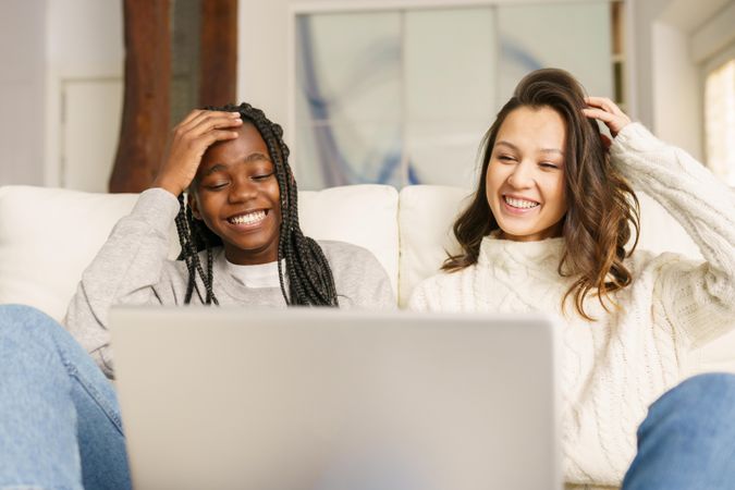 Two smiling women in jeans watching something together on a sofa on a pc