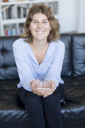 Front view portrait of a happy female holding a glass of water sitting on a couch in the living room at home