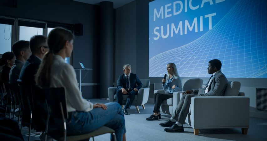 International Medical Summit: Host Asking Female Pharmaceutical CEO a Question In Front Of Audience Of Diverse Attendees.
