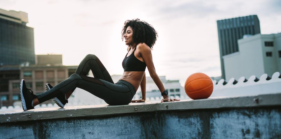 Woman in fitness wear sitting relaxed on rooftop fence with a basketball beside her