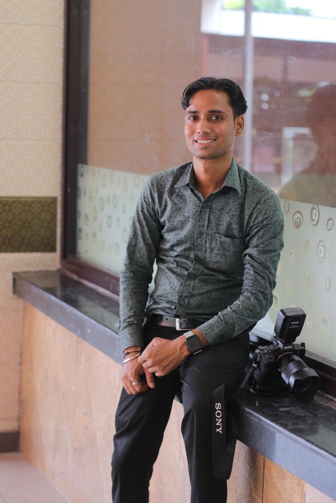 Man in gray dress shirt and dark pants sitting beside camera indoor