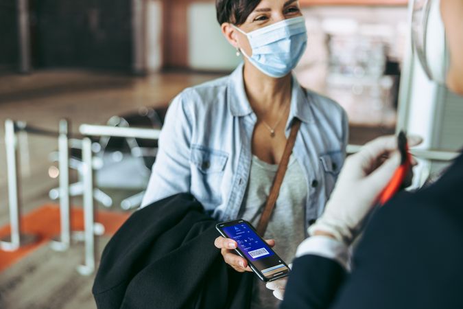Woman in face mask showing her boarding pass on mobile phone to flight attendant