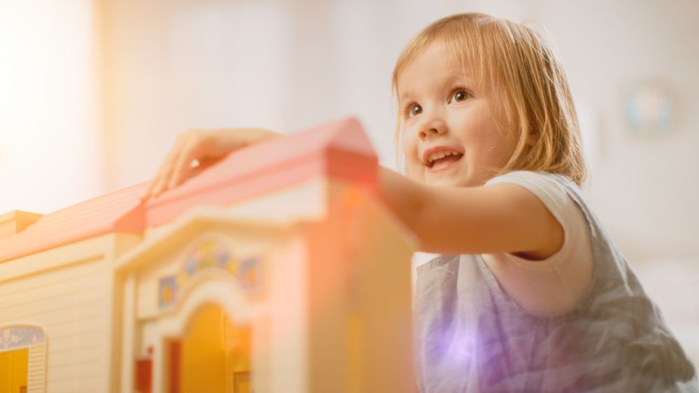 In the Children's Room: Happy Little Girl Playing with Doll House and Sitting on a Carpet.
