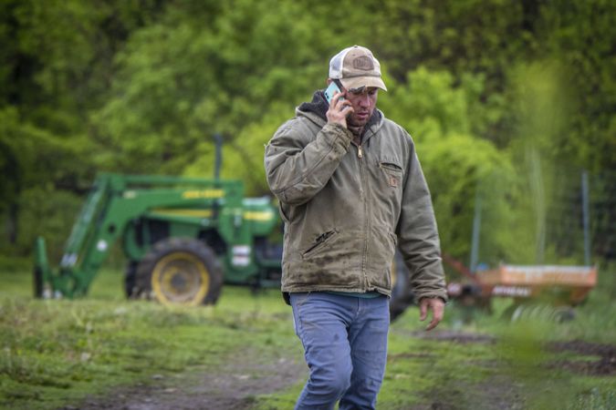 Copake, New York - May 19, 2022: Farmer walking away from tractor in field and talking on phone