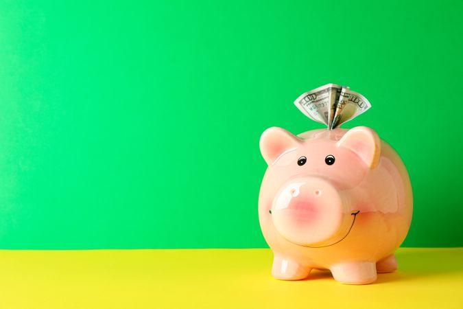 Happy piggy bank with one hundred dollar on color table against green background, space for text. Finance, saving money