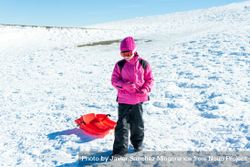Child in pink snow suit tugging red sled behind her up a snowy hill 5lJD6b